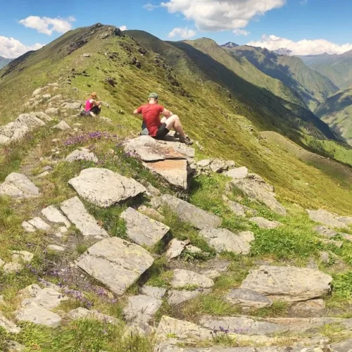 Panoramic view from the Unagira ridge in Tusheti, Georgia, featuring a hiker on a rocky outcrop overlooking steep green valleys and distant snowy Caucasus mountains under a sunny sky.