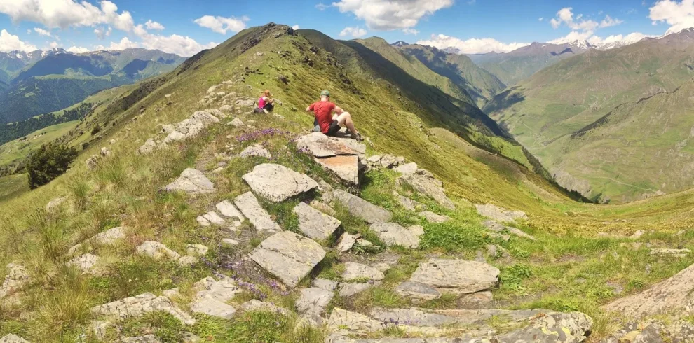 Panoramic view from the Unagira ridge in Tusheti, Georgia, featuring a hiker on a rocky outcrop overlooking steep green valleys and distant snowy Caucasus mountains under a sunny sky.