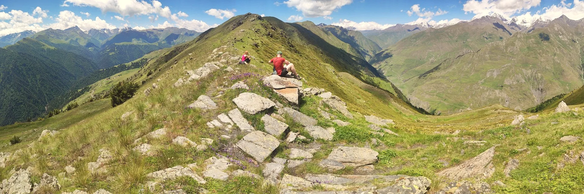 Panoramic view from the Unagira ridge in Tusheti, Georgia, featuring a hiker on a rocky outcrop overlooking steep green valleys and distant snowy Caucasus mountains under a sunny sky.