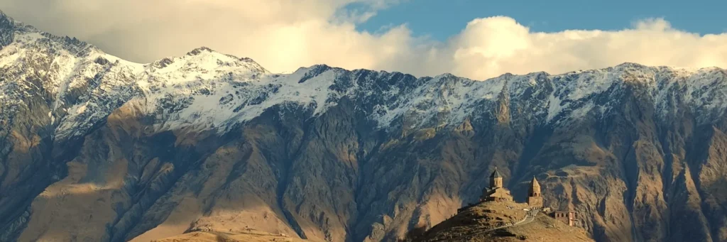 Panoramic view of Gergeti Trinity Church perched on a hill with rugged snow-dusted Caucasus mountain peaks and blue sky in Kazbegi, Georgia.