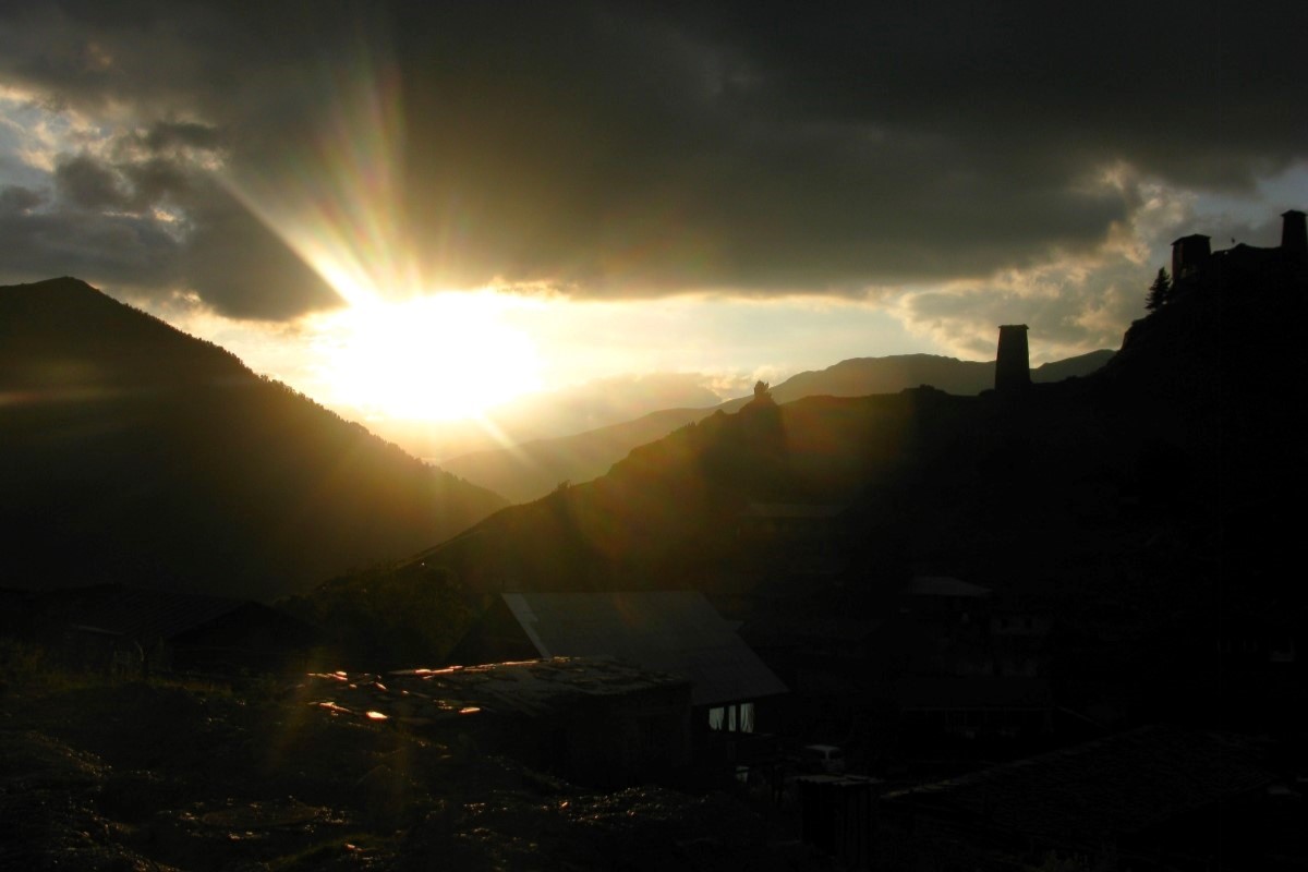 Dramatic golden hour sunset rays breaking through dark mountain clouds behind medieval stone defense towers in Tusheti