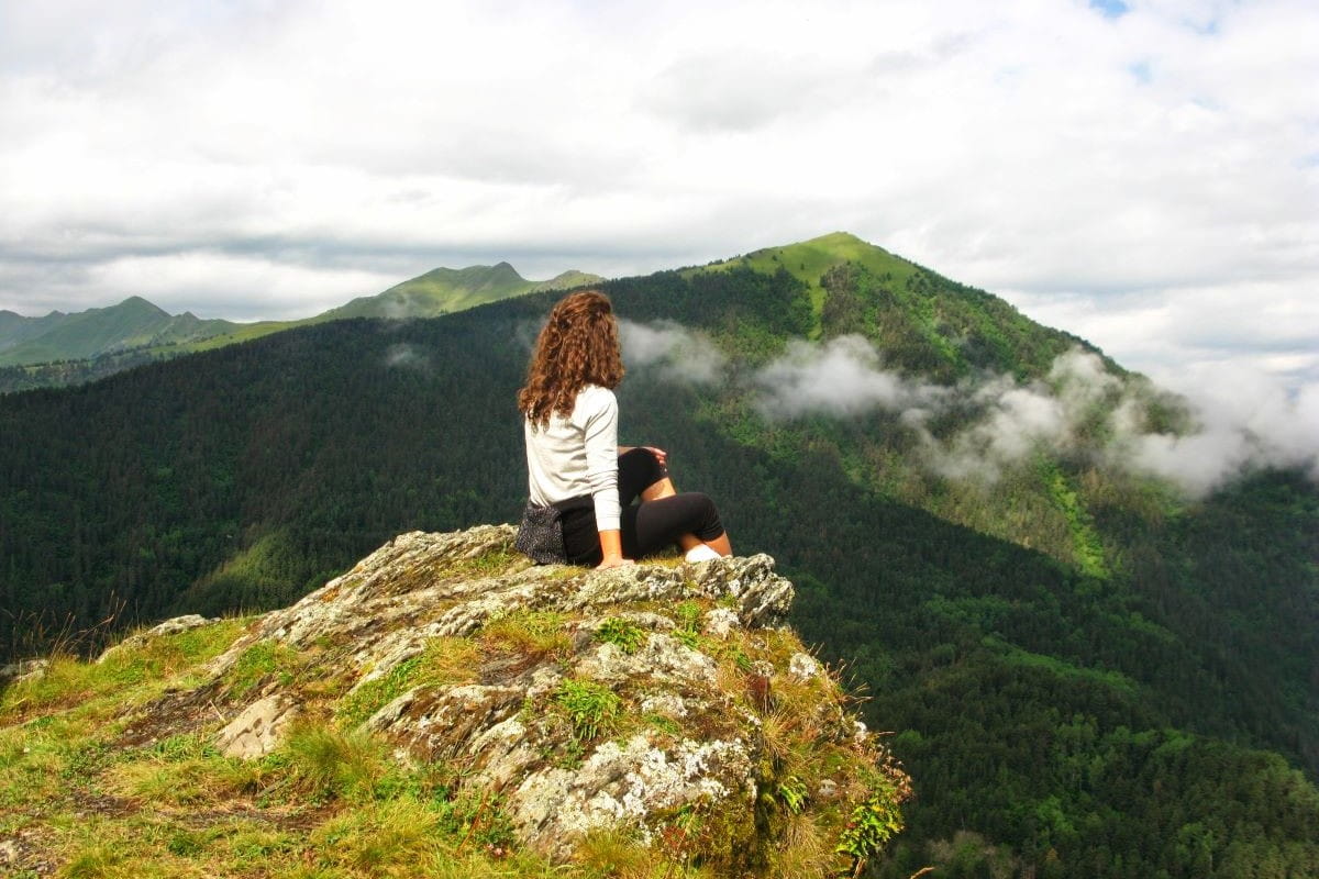 Hiker sitting on a high rock viewpoint overlooking pine forests and alpine clouds in Tusheti Georgia